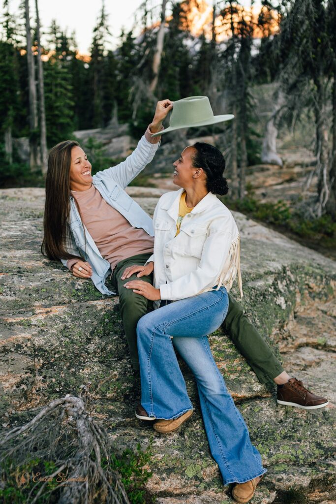 A woman lifts off the hat of another while they sit on a large boulder in the North Cascades during their Washington couples session. 