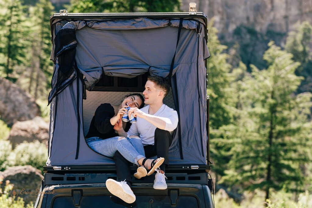 An adventure session couple sits in a roof top tent while sharing beers. 