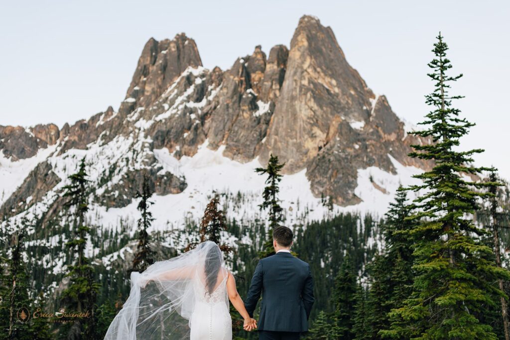 A bride lifts her wedding veil while holding her groom's hand as they admire the North Cascades during their elopement. 