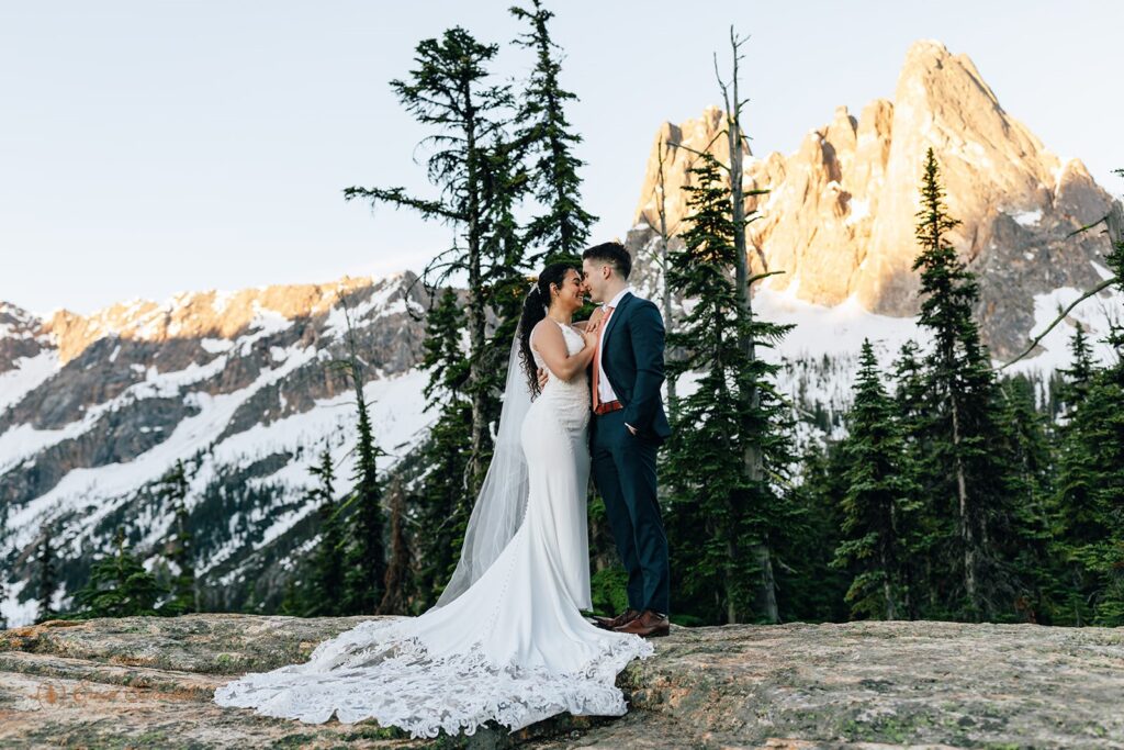 A North Cascades elopement couple embraces as the sun illuminates mountain peaks in the distance. 