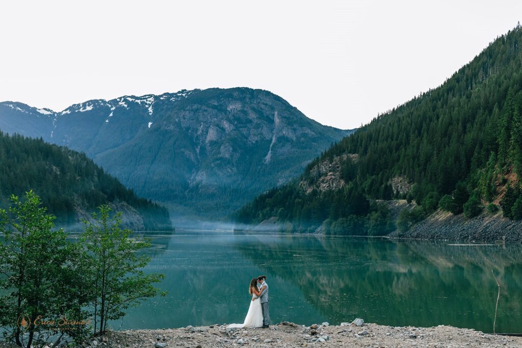 A couple wearing formal wedding attire embraces on the shores of Diablo Lake during their North Cascades elopement