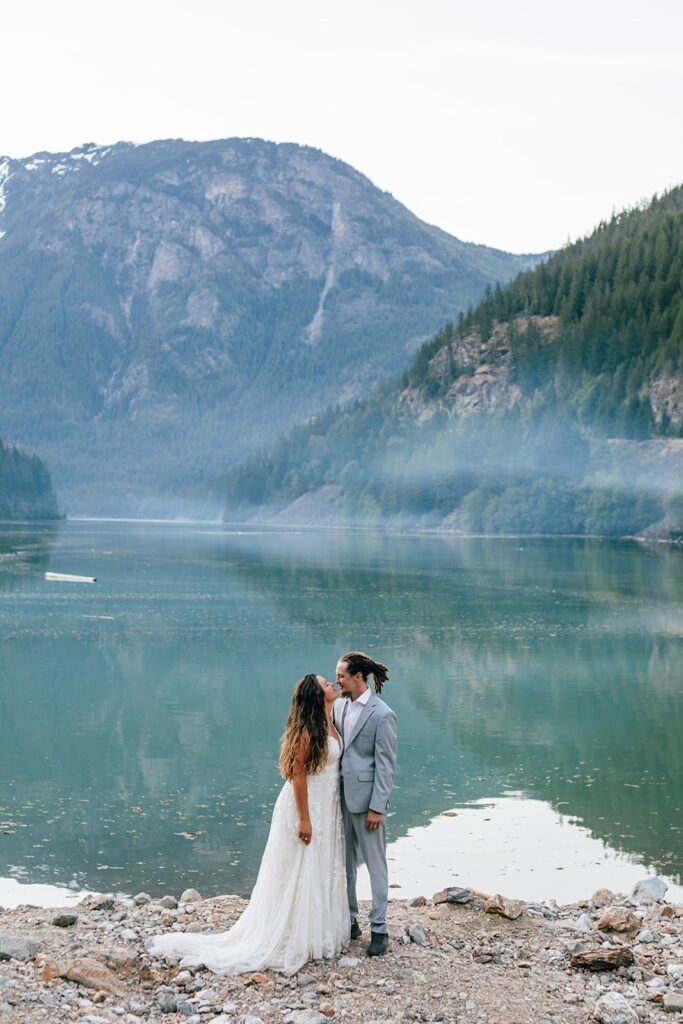 A groom in a grey wedding suit admires his bride, who is wearing a long wedding dress, on the shores of Diablo Lake. 