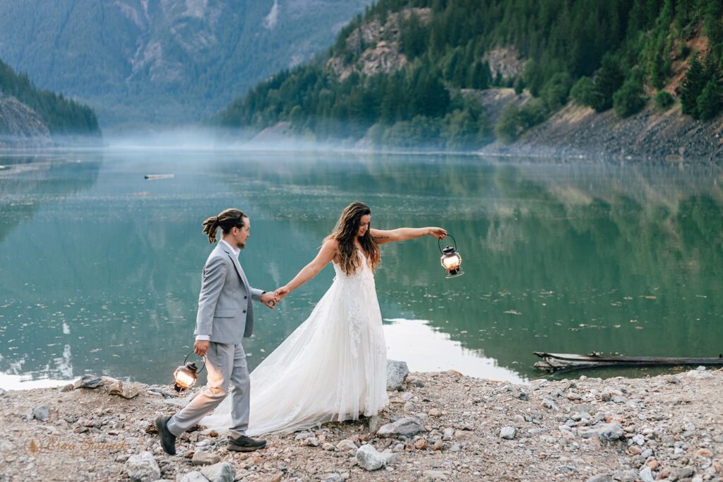 A groom in a grey wedding suit is lead by his bride, who is wearing a long wedding dress and holding a lantern,  as they walk along Diablo Lake. 
