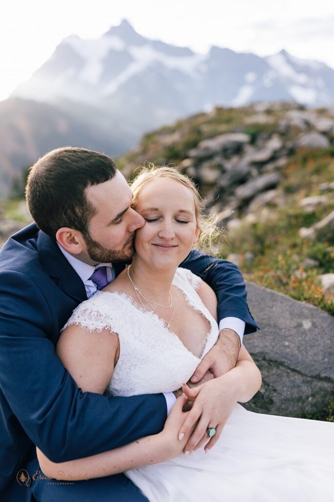 A groom in a navy suit sits with his bride and embraces her during their Sunrise Artist Point elopement. 