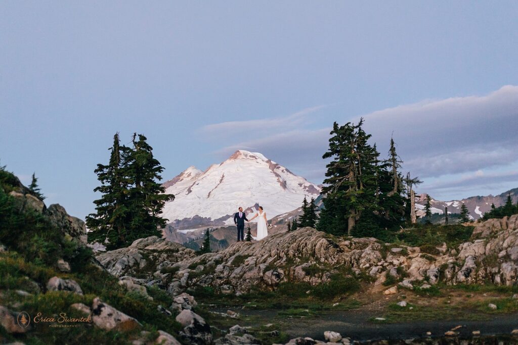 A North Cascades elopement couple stands long a cliff in the distance with Mt. Baker in the background. 