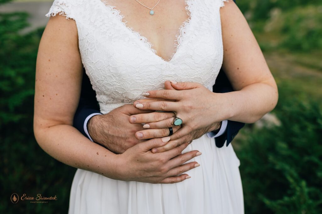 A bride in an a-line, white wedding gown holds her groom's hands that are wrapped around her waist. 