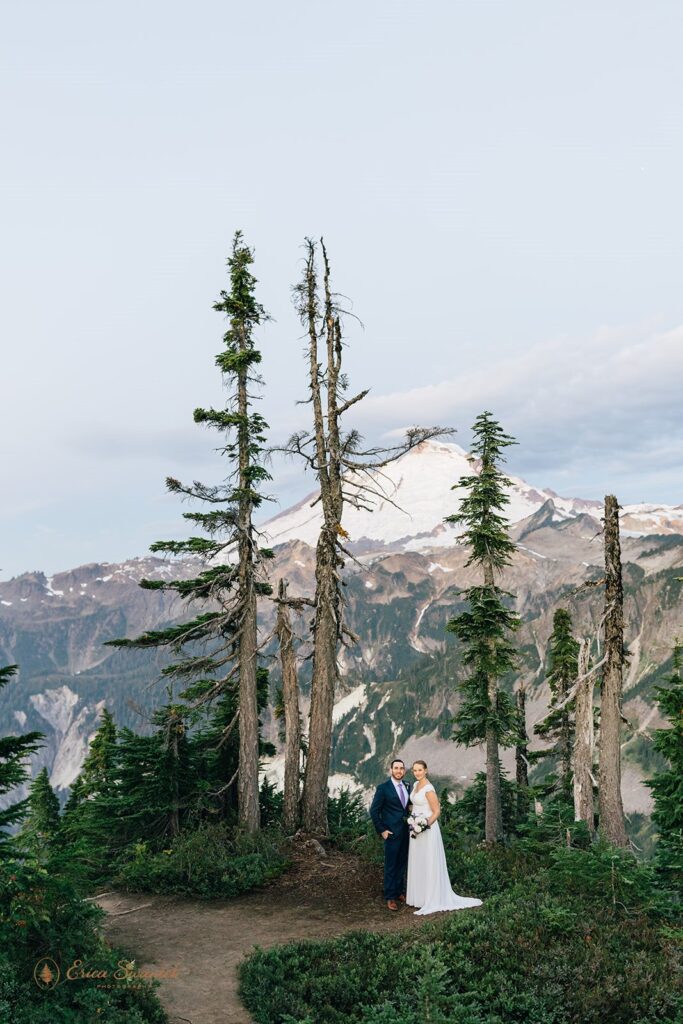 A groom dressed in a navy suit stands with his bride, who is dressed in a long wedding gown, in front of a snowy mountain during their Artist Point elopement. 