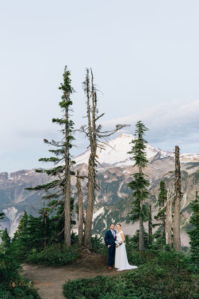 A couple poses for a smiling wedding portrait in front of a snowy Mt. Baker during their Sunrise elopement at Artist Point. 