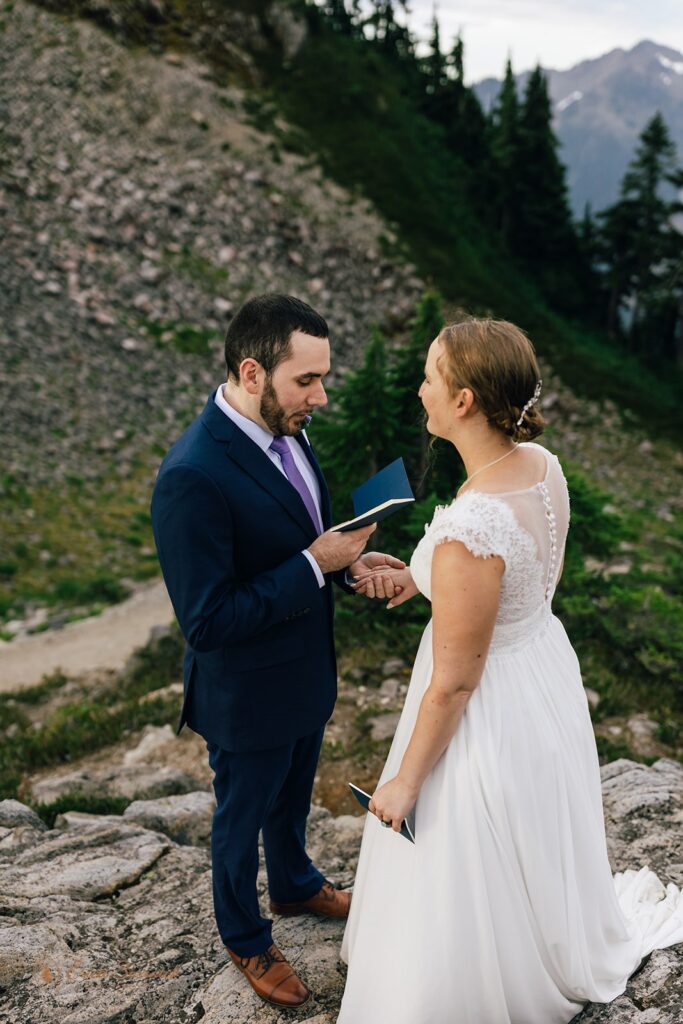 A groom in a navy suit, brown oxford shoes, and a purple tie holds a blue vow book and his bride's hand during their Artist Point ceremony. 