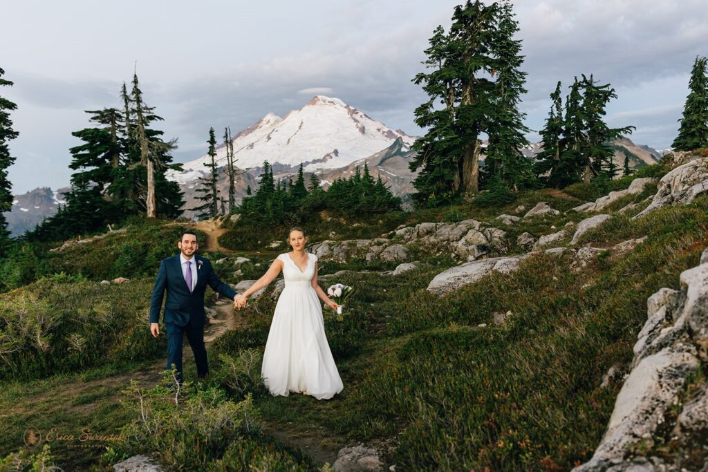 A groom wearing a navy suit and purple tie holds his bride's hands, who is wearing an a-line wedding dress in a meadow in Washington. 
