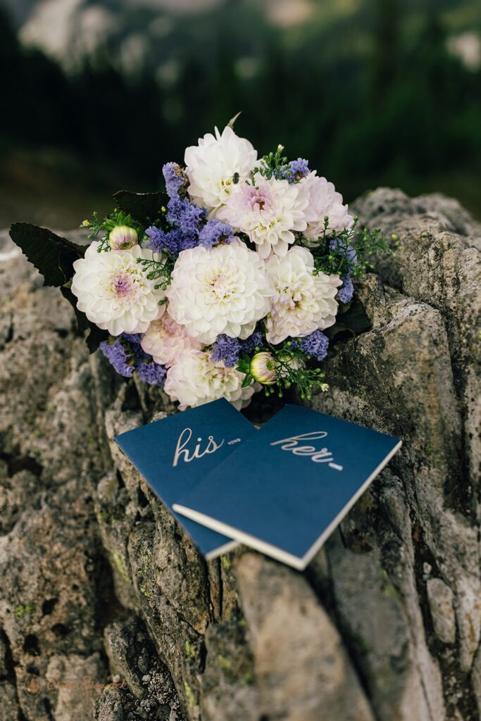 A bright floral bridal bouquet with purple and pink flowers near navy blue vow books. 