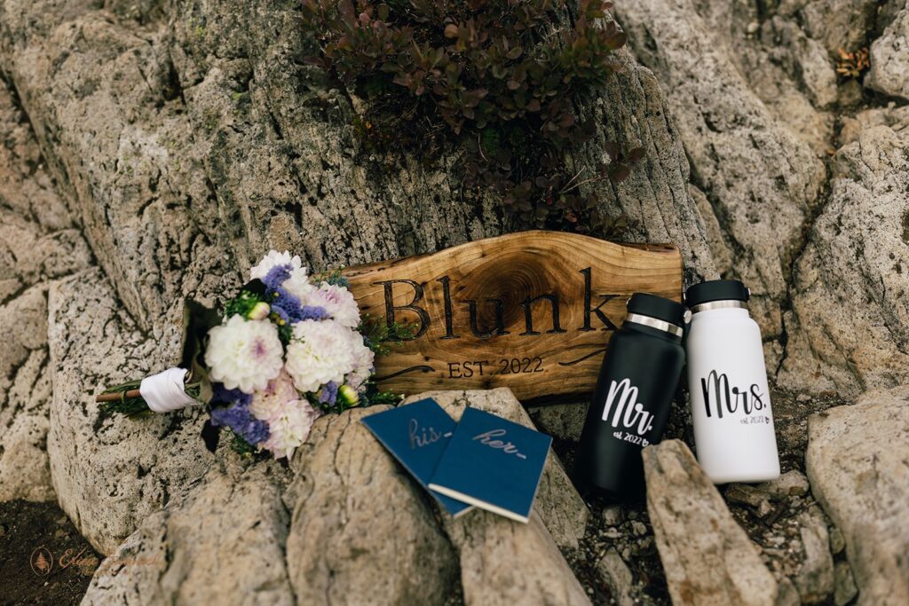 A sign reading Blunk rests alongside a rock, along with a black Mr. water bottle, white Mrs. water bottle, navy vow books and a bright bridal bouquet with purple and pinks flowers. 