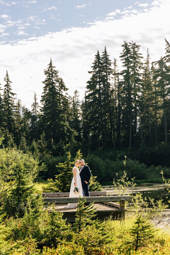 A couple in formal wedding attire stands on a bridge in an evergreen forest during their North Cascades elopement.