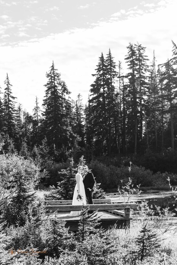 A North Cascades elopement couple stands on a bridge in an evergreen forest taking Washington wedding portraits. 
