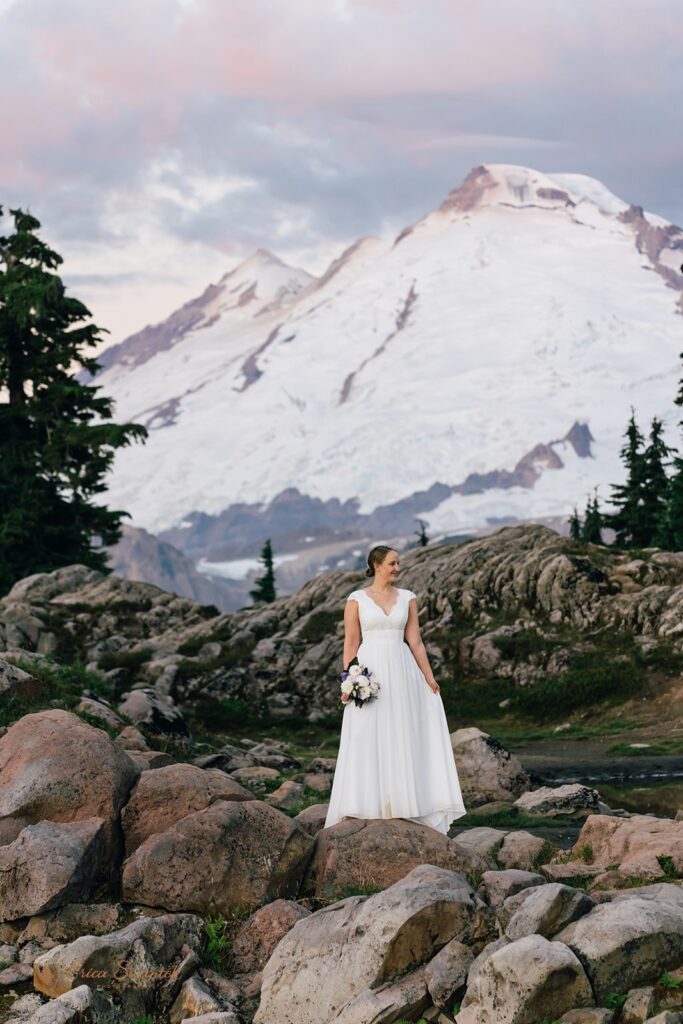A bride, wearing a a-line, floor length white wedding gown, stands in front of Mt. Baker while holding a rose bridal bouquet. 