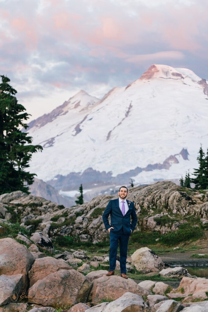 A groom, wearing a navy suit, brown shoes and a purple tie, stands in front of Mt. Baker. 