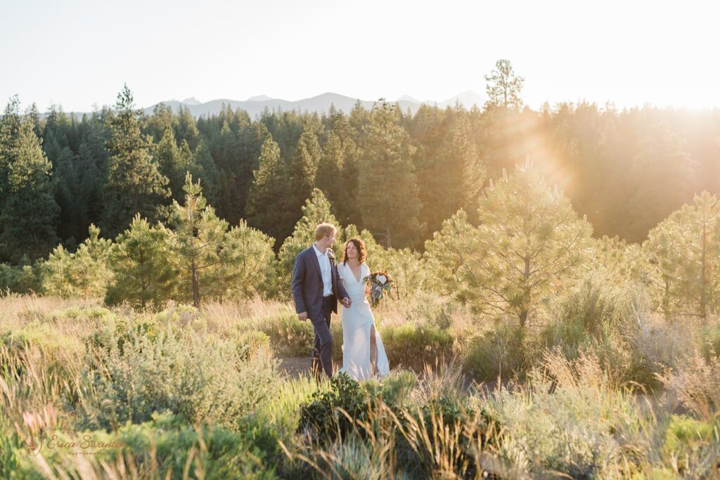 An Oregon intimate wedding couple walks through a meadow near an evergreen forest at Shevlin Park. 