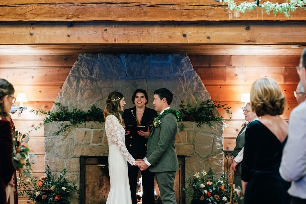 An Oregon officiant conducts an intimate wedding ceremony near a fireplace decorated with greenery and floral arrangements. 