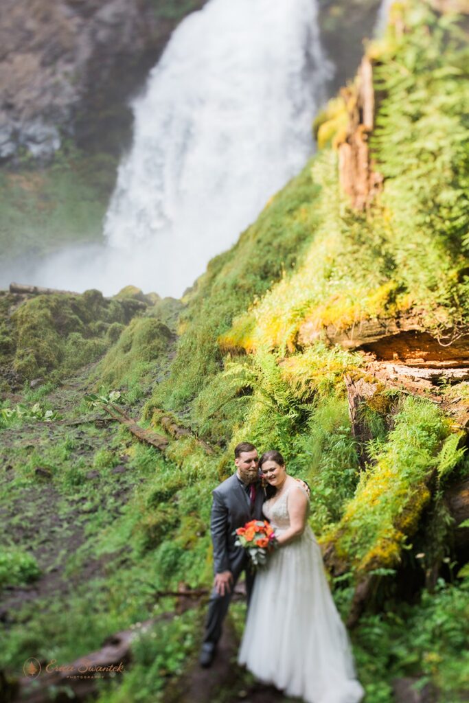 A groom in a grey wedding suit embraces his bride, who is holding an orange bridal bouquet, near Sahalie Falls.