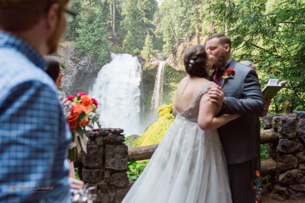 A bride in a long, white wedding gown kisses her groom near Sahalie Falls during their Oregon elopement ceremony. 