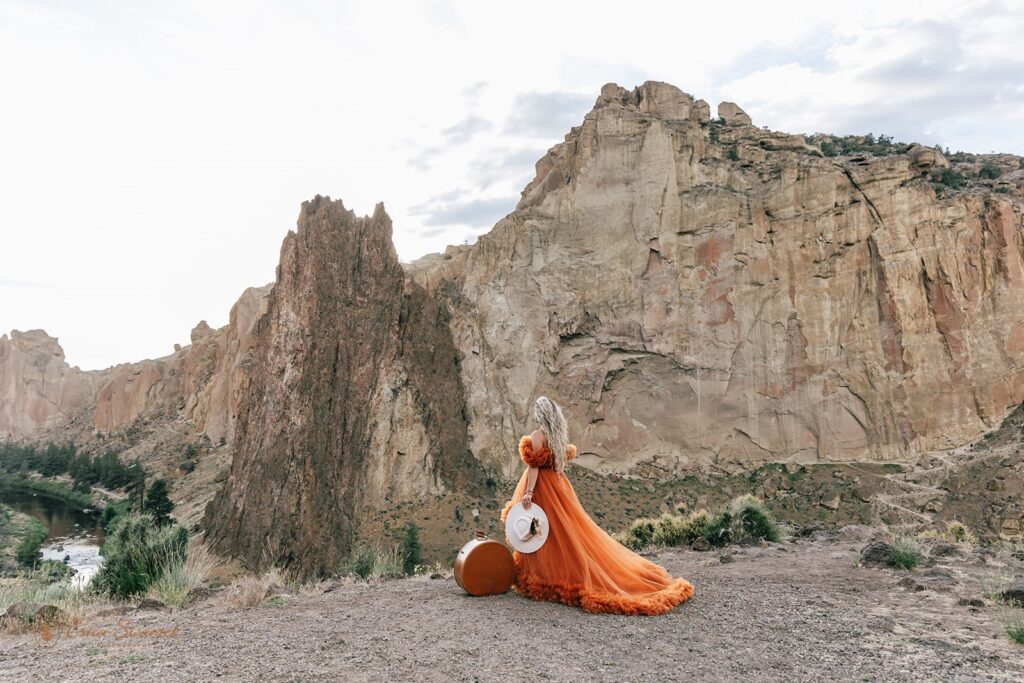 A bride in a boho orange dress embraces admires the rock formations of Rock Smith State Park while standing next to a piece of round, vintage luggage. 