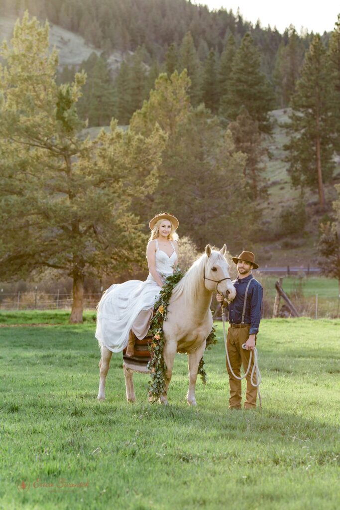 A bride in a long, white wedding dress and cowboy hat sits on a horse while her husband holds the reigns at Wine Down Ranch