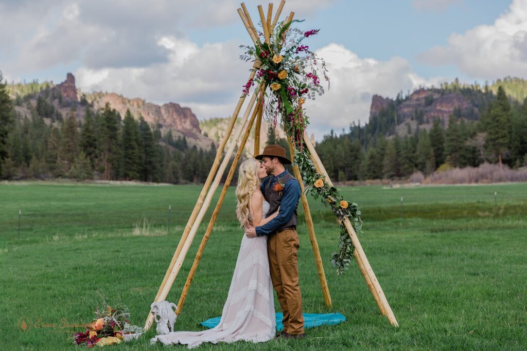 A bride and groom kiss beneath a bohemian ceremony arch during their intimate wedding at Wine Down Ranch. 