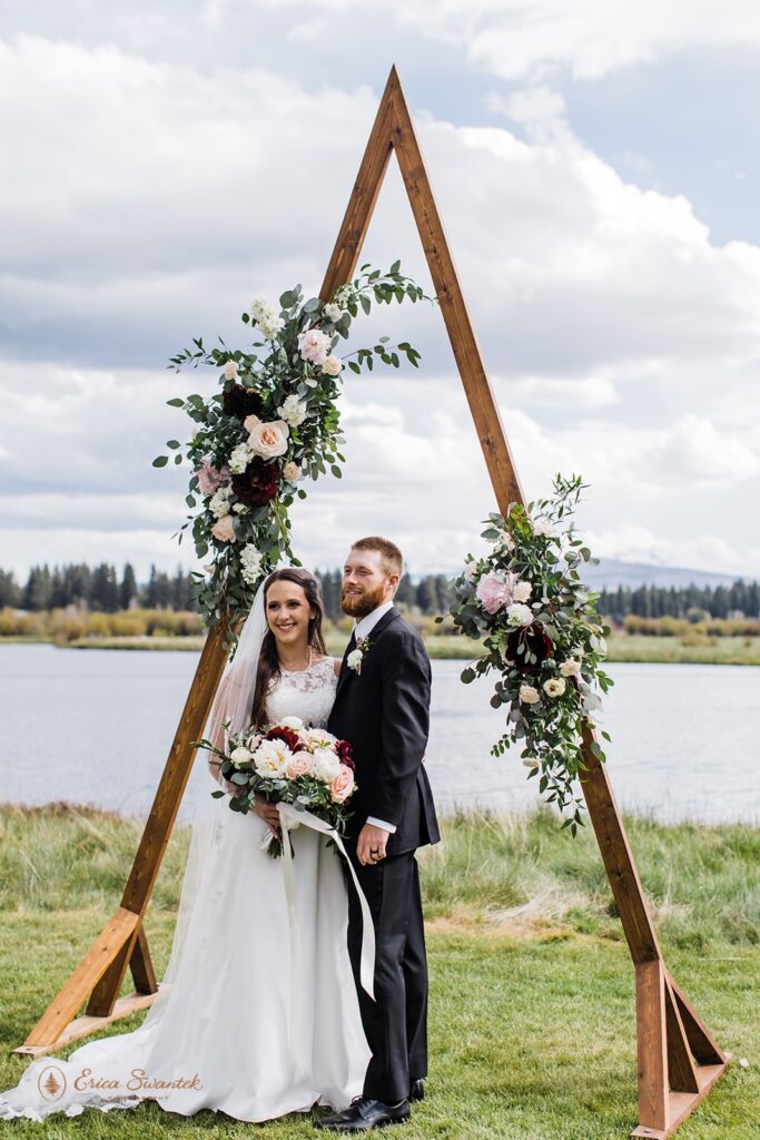A bride holds a burgundy, pink and white rose bridal bouquet while embracing her groom at beneath an a-frame wedding arch at Black Butte Ranch in Sisters, Oregon. 