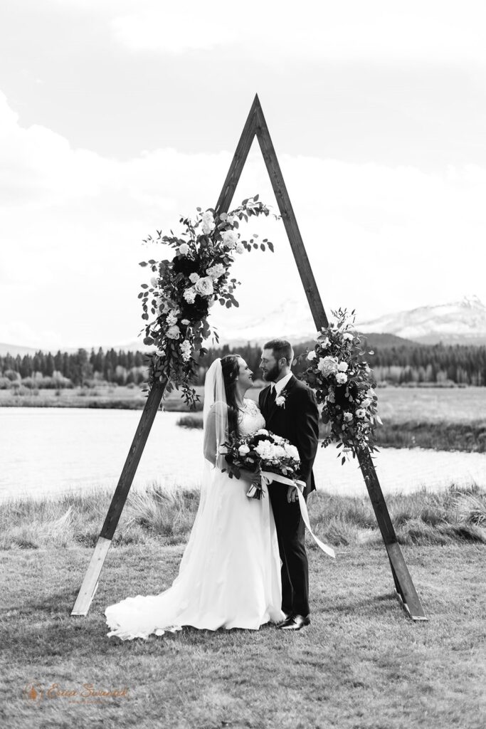 A couple dressed in formal wedding admire one another while embracing beneath a floral decorated a-frame arch at Black Butte Ranch in Sisters, Oregon. 