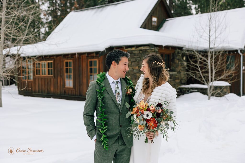 A groom in a forest green suit and a bride in a long wedding dress and fur stole embrace in front of Lake Creek Lodge in Winter. 
