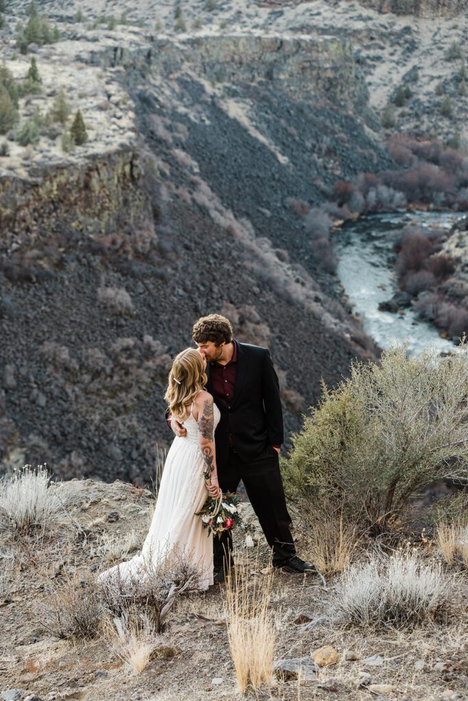 A bride and groom kiss on a hillside overlooking a canyon during their Bend, Oregon, elopement. 