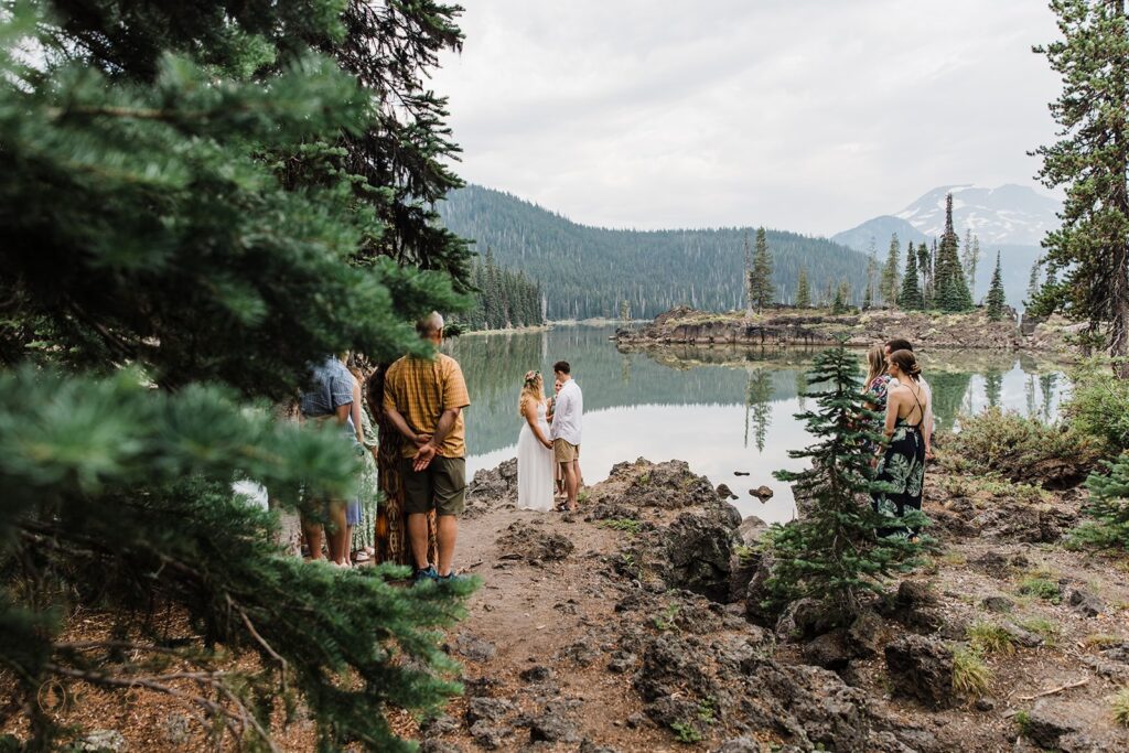 A Sparks Lake elopement couple holds hands during a lakeside vow ceremony with family and friends near Bend, Oregon. 