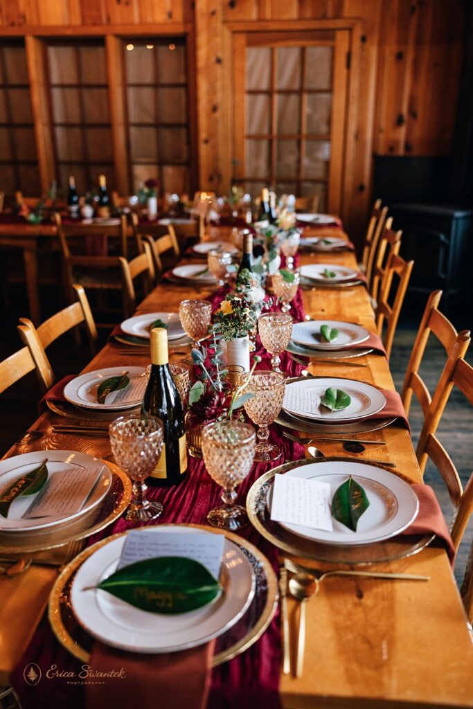 A burgundy tablescape on a wooden table accented with green leaf place card and crystal glasses. 