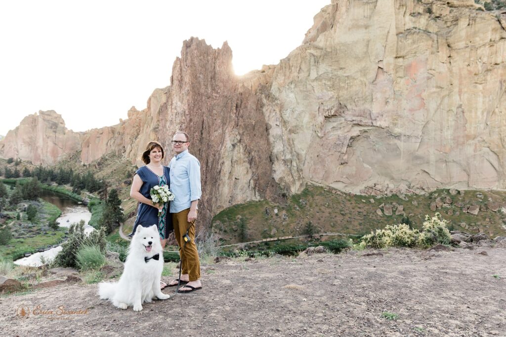 A Smith Rock State Park elopement couple poses for a couples portrait with their dog, who is wearing a black bow tie. 