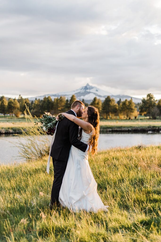 A Bend, Oregon, elopement couple embraces in a meadow alongside a river with a snowy mountain peak in the distance. 
