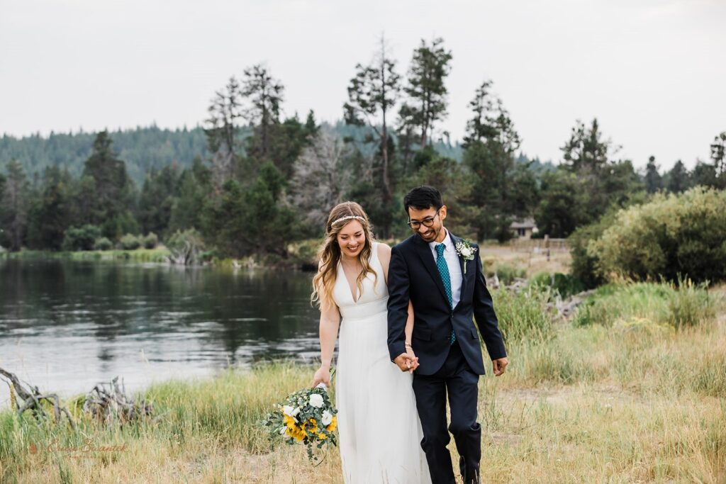 A bride in in a floor length wedding dress holds a sunflower bouquet while walking with her groom along the banks of Sunriver at Sunriver Resort. 