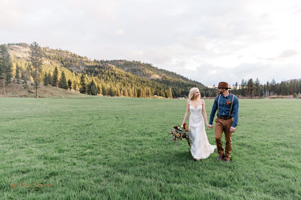 A boho elopement couple hold hands while walking in a meadow at Wine Down Ranch. 