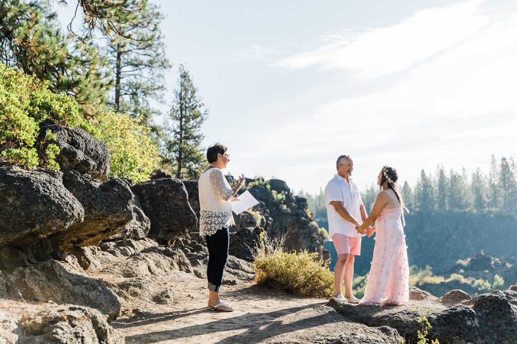 An Oregon elopement couple, dressed in matching pink wedding attire, recites vows with an officiant at Tumalo Falls. 