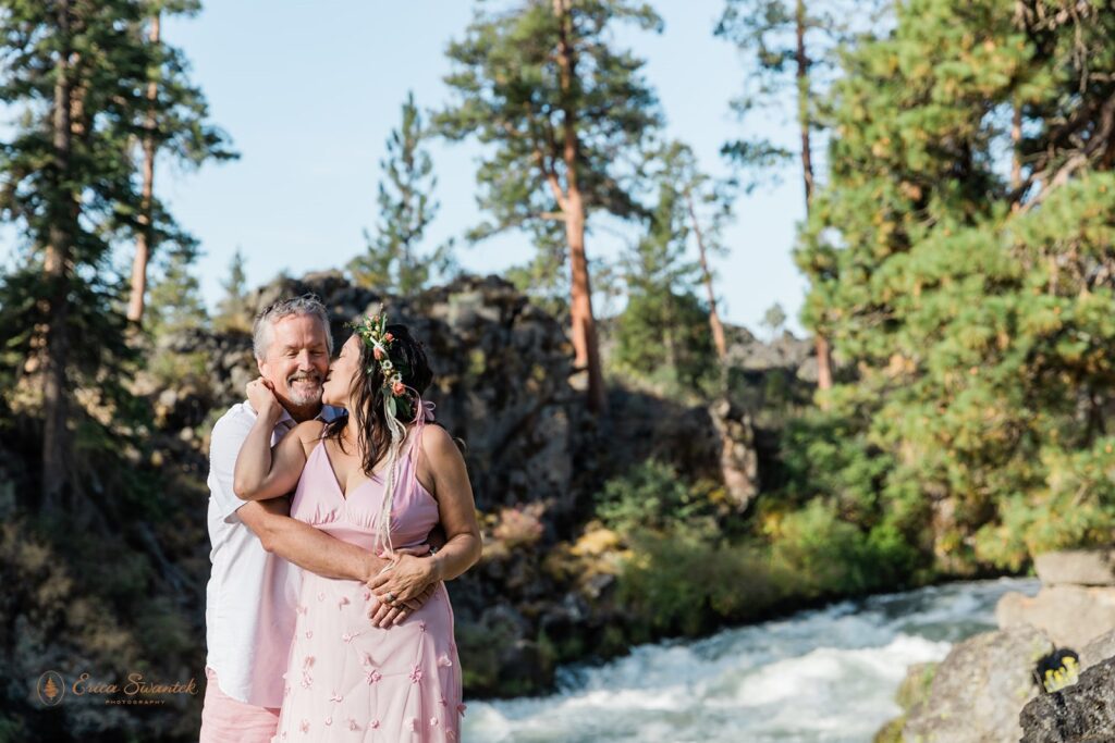 An Tumalo Falls elopement couple, dressed in matching pink wedding attire, kiss near the waterfall in Oregon. 