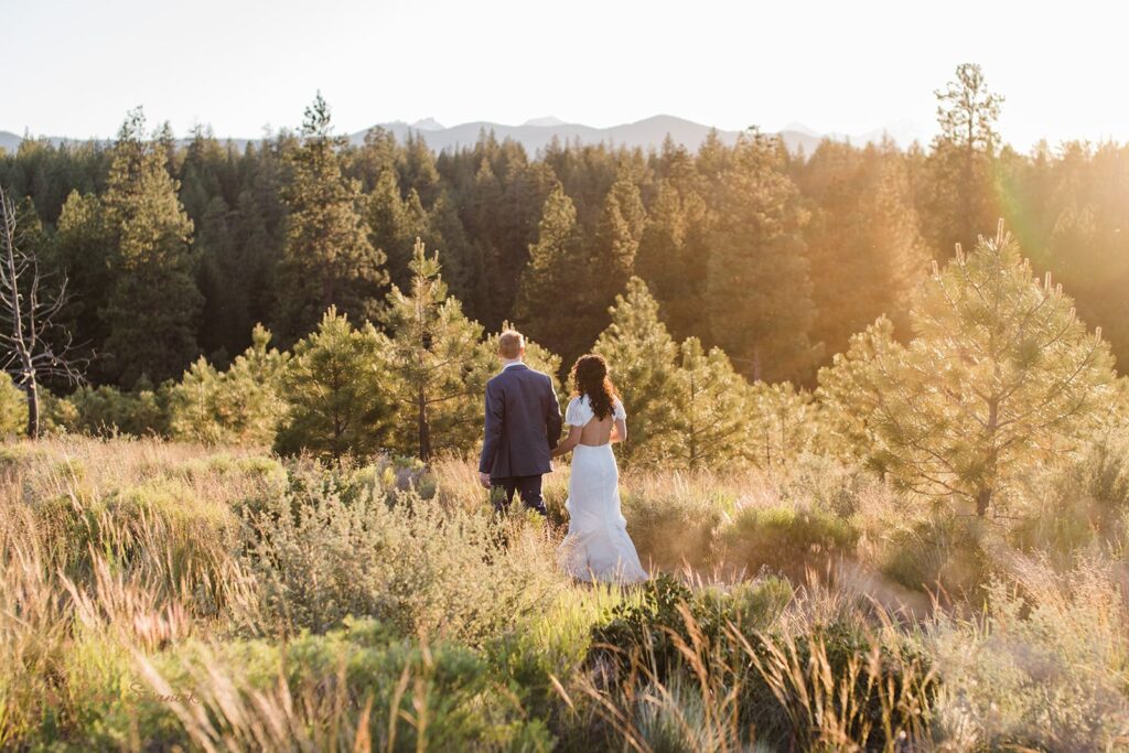 A couple dressed in formal wedding attire hold hands while walking through a meadow during golden hour at Shevlin Park. 