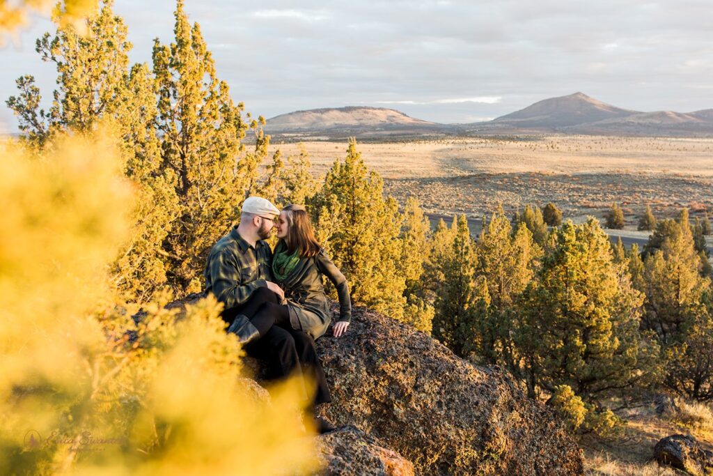 An adventure elopement couple sits atop a rock surrounded by evergreen trees that overlooks Crooked River Grasslands at Sunset. 