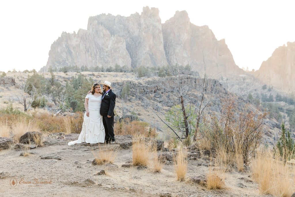 An adventure elopement couple dressed in formal wedding attire embraces at Sunset at Smith Rock State Park. 