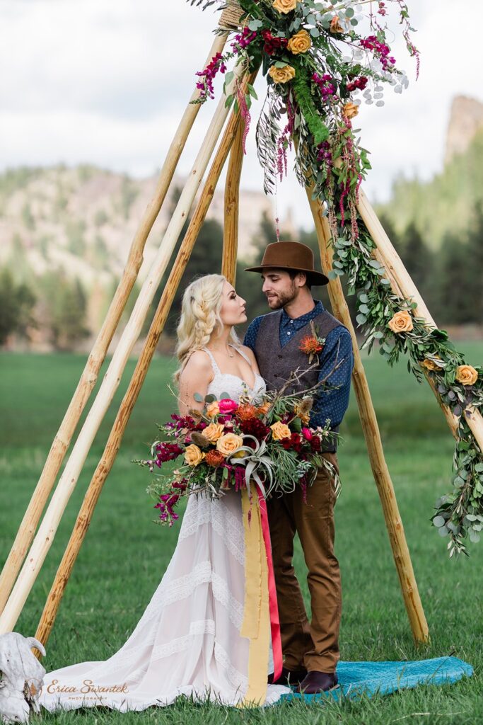 A boho elopement couple embraces beneath a teepee arch decorated with burgundy and apricot roses at Wine Down Ranch. 