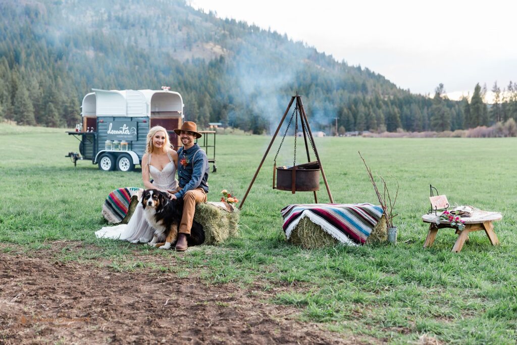 A boho elopement couple sits with their dog on a hay bale covered with a blanket near a campfire at Wine Down Ranch near Bend. 