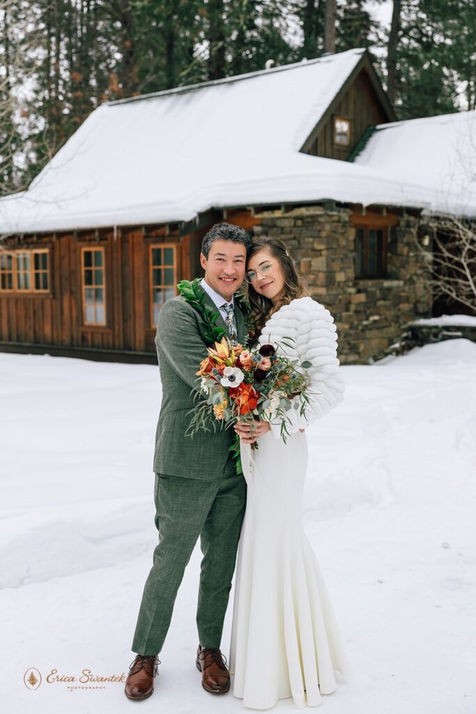 A bride dressed in a floor length wedding dress and white fur bridal wedding shawl holds a bright bridal bouquet while standing by her groom, who is wearing a forest green suit and Maile Lei. 