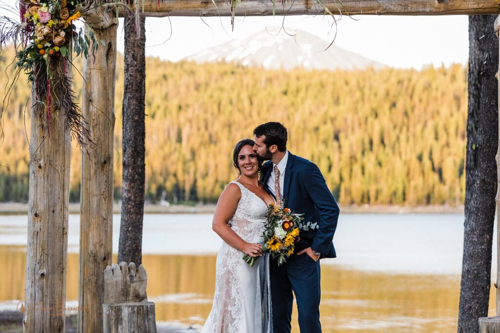 A groom kisses his bride beneath a wooden arch at Elk Lake Lodge, an intimate Bend wedding location. 