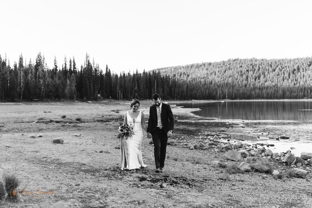An intimate Oregon wedding couple dressed in formal ceremony attire holds hands while walking along the shore of Elk Lake. 