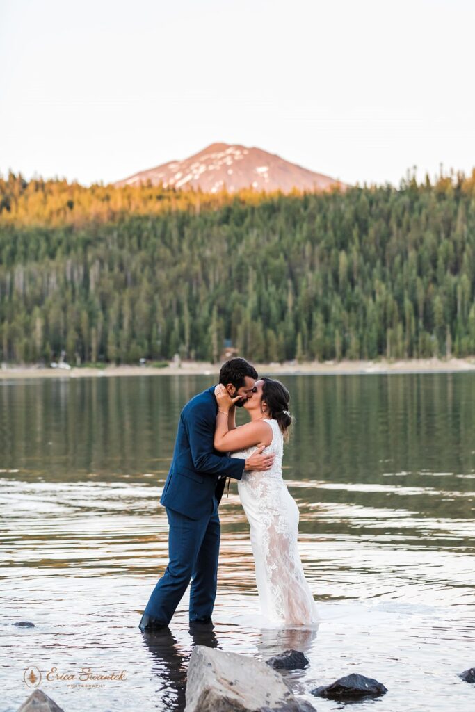 A groom in a navy suit and a bride in a long, lace wedding dress are kissing while standing in Elk Lake at Sunset. 