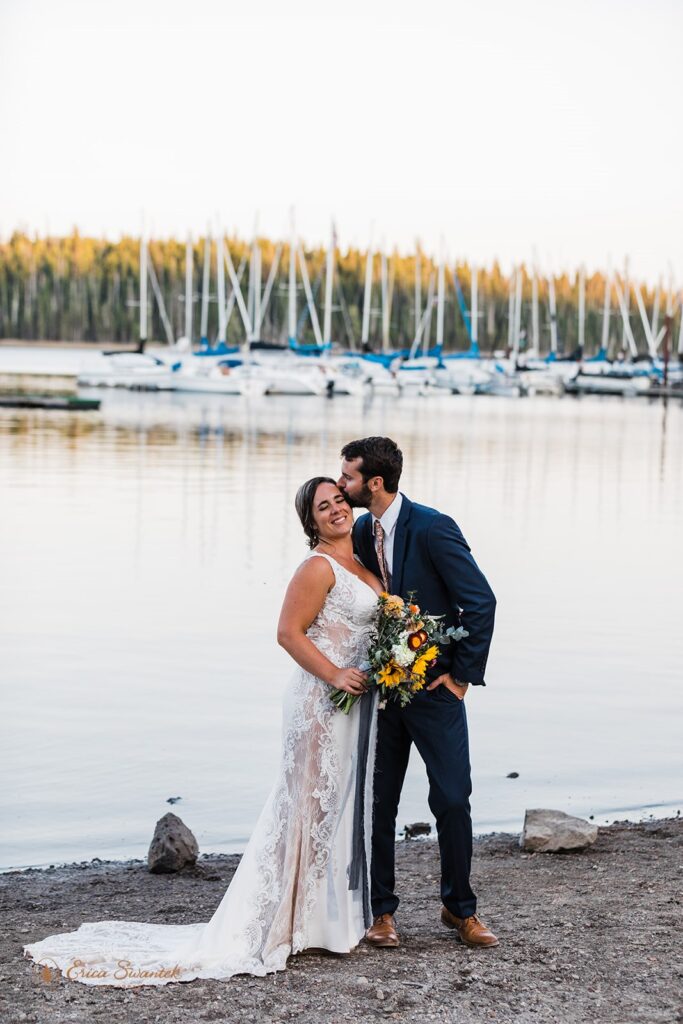 A groom kisses his bride on the shores of Elk Lake in Bend, Oregon, with sail boats in the background. 
