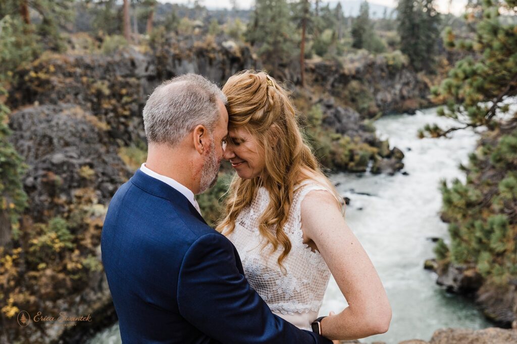 A bride and groom dressed in formal wedding attire embrace near Dillon Falls, an intimate wedding location in Bend, Oregon. 